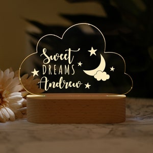 Peut inclure: Une veilleuse en forme de nuage en acrylique blanc avec le texte "Sweet Dreams Andrew", un croissant de lune et des étoiles. La lumière est posée sur une base en bois.
