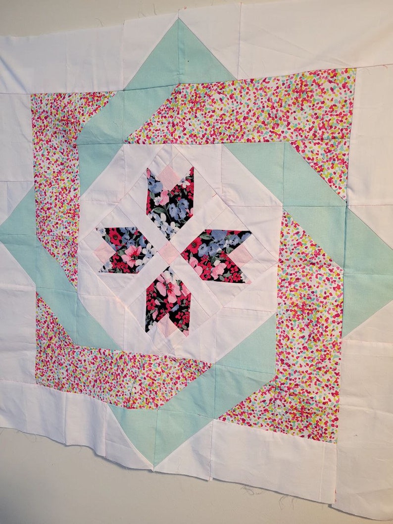 Pode incluir: Um edredom branco com um padr&atilde;o floral no centro. O edredom &eacute; feito de quadrados e tri&acirc;ngulos em tons de rosa, branco e verde.