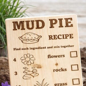 May include: Wooden sign with a "Mud Pie Recipe" engraved on it. The recipe includes flowers, rocks, grass, and dirt. The sign is on a wooden surface with a spoon of soil nearby.