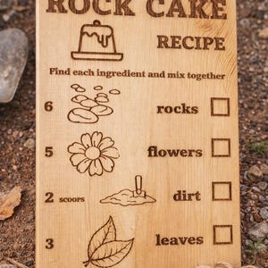 Pu&ograve; includere: Cartello in legno "Ricetta Torta di Roccia" con testo e illustrazioni incise. Include un elenco di ingredienti: rocce, fiori, terra e foglie. Presenta un'etichetta "Handmade" e uno striscione "Bestseller".