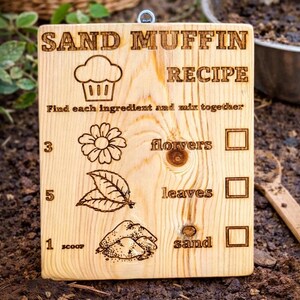 Pu&ograve; includere: Cartello in legno con ricetta per muffin di sabbia incisa. Include illustrazioni di cupcake, fiore, foglie e sabbia, con caselle di controllo. Il testo recita: "Trova ogni ingrediente e mescola insieme".