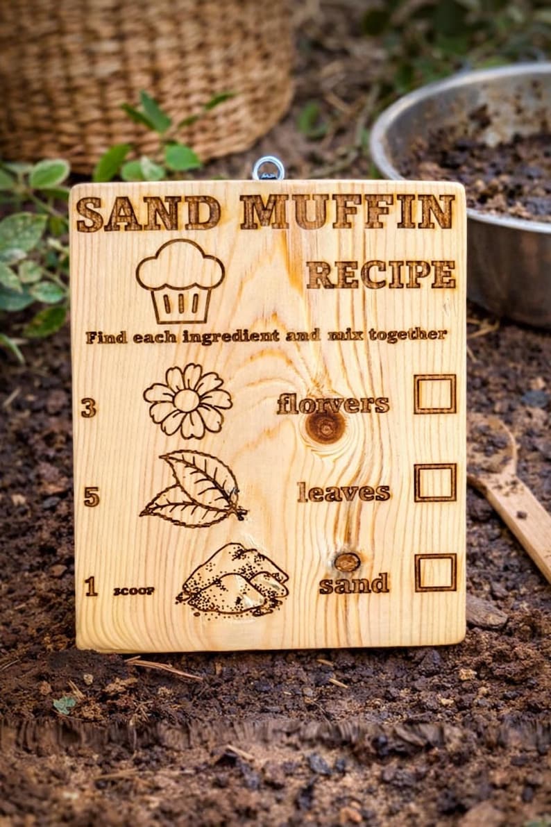 Pu&ograve; includere: Cartello in legno con ricetta per muffin di sabbia incisa. Include illustrazioni di cupcake, fiore, foglie e sabbia, con caselle di controllo. Il testo recita: "Trova ogni ingrediente e mescola insieme".
