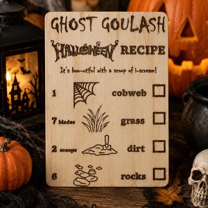 Pu&ograve; includere: Un cartello di ricetta di Halloween in legno con la scritta "Ghost Goulash Halloween Recipe". Il cartello include illustrazioni di una ragnatela, erba, terra e rocce. Sono visibili anche una piccola zucca, un teschio e un ragno.