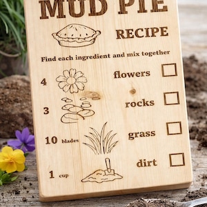 Puede incluir: Cartel de madera con la "Receta de pastel de barro" grabada. La receta incluye flores, rocas, hierba y tierra. El cartel está sobre una superficie de madera con una cuchara de tierra cerca.