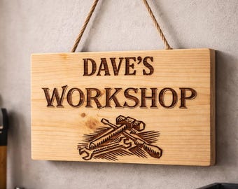 May include: A rectangular light-colored wooden sign hanging from a jute rope. The sign features the engraved text DAVE'S WORKSHOP in a bold font, positioned above an engraved graphic illustration of a hammer, a saw, and two wrenches.