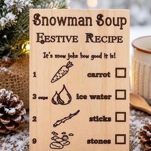 Pu&ograve; includere: Targa in legno con la scritta "Snowman Soup Festive Recipe". La ricetta include carote, acqua ghiacciata, bastoncini e pietre. La targa ha un design rustico inciso, perfetto per l'arredamento invernale o festivo.