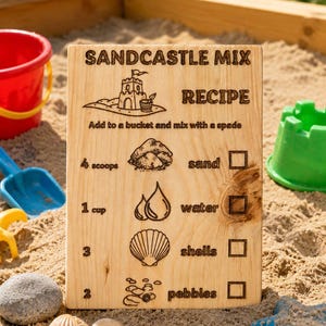 Pu&ograve; includere: Un cartello di legno con il testo "SANDCASTLE MIX RECIPE" e le istruzioni per fare castelli di sabbia. Il cartello include illustrazioni di sabbia, acqua, conchiglie e ciottoli. Un secchio rosso, una pala blu e un secchio verde sono sullo sfondo.