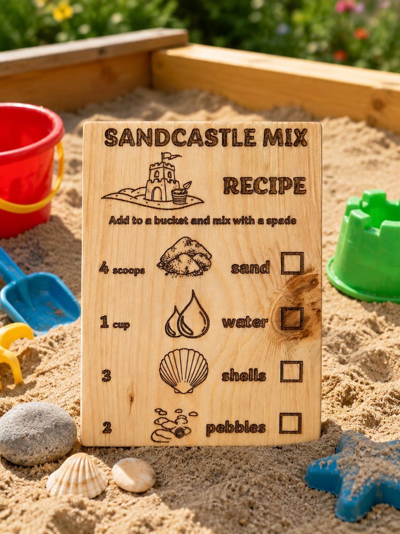 Pu&ograve; includere: Un cartello di legno con il testo "SANDCASTLE MIX RECIPE" e le istruzioni per fare castelli di sabbia. Il cartello include illustrazioni di sabbia, acqua, conchiglie e ciottoli. Un secchio rosso, una pala blu e un secchio verde sono sullo sfondo.