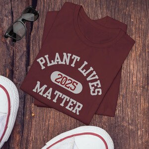 May include: A maroon t-shirt with white text that reads "Plant Lives Matter 2025". The text is in a collegiate font.