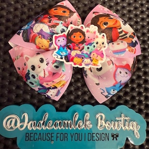 May include: A large pink hair bow with cartoon characters and a central sticker featuring a group of characters. The bow has a repeating pattern of cartoon characters. The text "@Jadeamlek Boutiq" and "BECAUSE FOR YOU I DESIGN" is visible on a blue background.