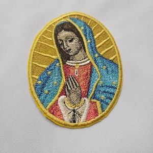 Puede incluir: Parche bordado con una representación de la Virgen María con una túnica azul y roja, con un halo dorado y un fondo amarillo.