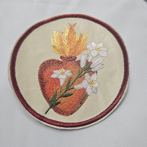 May include: Embroidered patch featuring a heart with flames, a floral sprig, and a cream-colored background. The heart is in shades of red and orange, with yellow and orange flames. The patch is round, with a burgundy border.