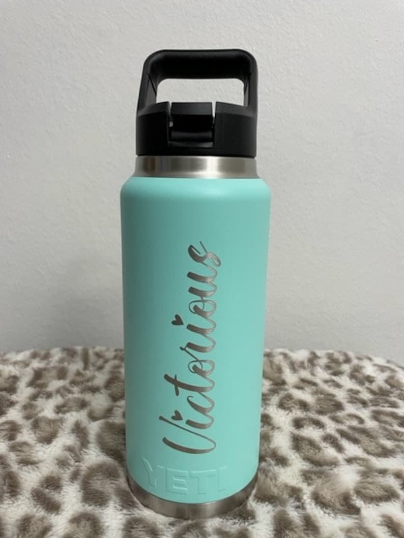 Puede incluir: Una botella de agua Yeti azul turquesa con tapa y asa negras. La botella tiene la palabra "Victorious" grabada en el lateral en una fuente plateada.