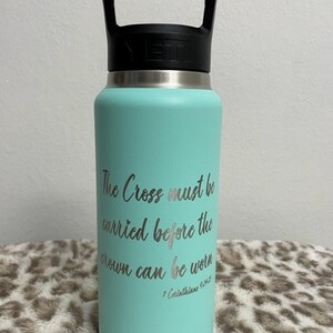 Puede incluir: Una botella de agua YETI turquesa con una manija negra y una inscripci&oacute;n plateada que dice "The Cross must be carried before the crown can be worn 1 Corinthians 9:25".