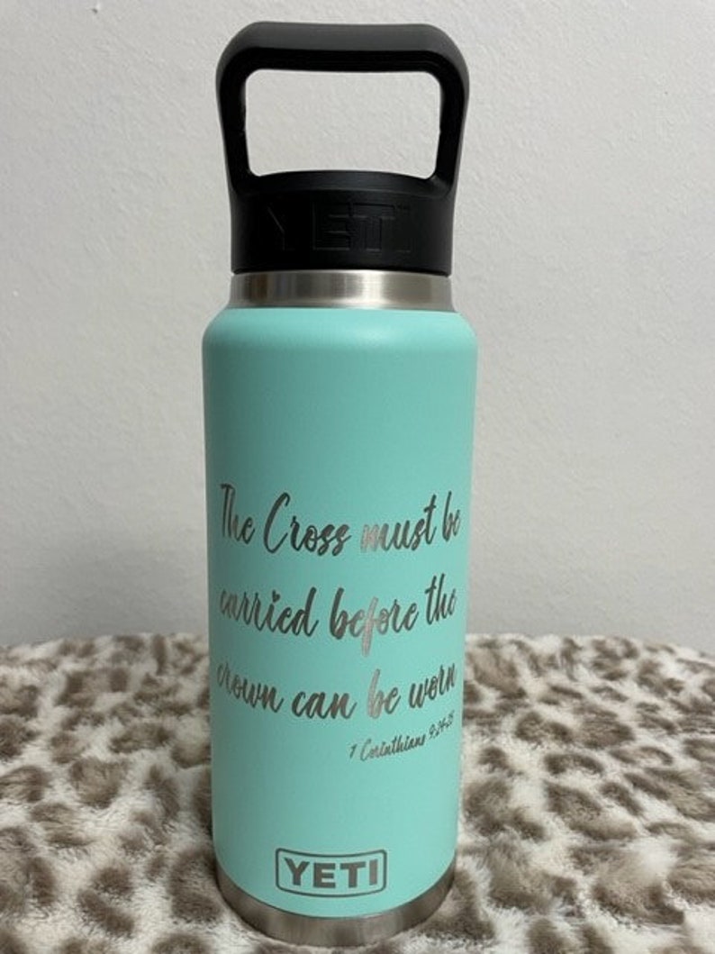 Puede incluir: Una botella de agua YETI turquesa con una manija negra y una inscripci&oacute;n plateada que dice "The Cross must be carried before the crown can be worn 1 Corinthians 9:25".