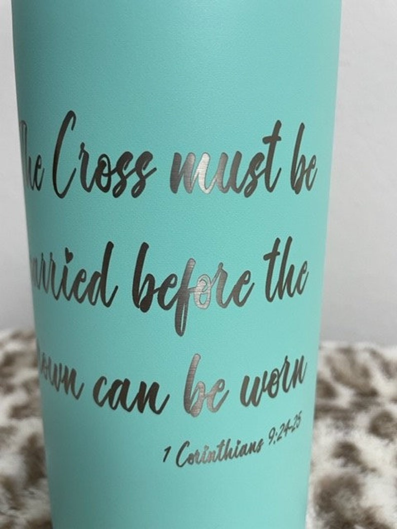 Puede incluir: Un vaso azul turquesa con una inscripci&oacute;n plateada que dice "The Cross must be carried before the crown can be worn 1 Corinthians 9:24-25".