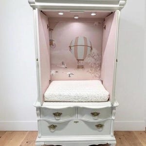 May include: A white wooden armoire featuring two lower drawers with ornate brass pulls. The open upper section reveals a soft pink interior backdrop with a hot air balloon and swan illustration, illuminated by three small lights above a white cushion.