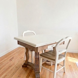 May include: A square dining table with a white top and a natural wood base featuring a small drawer. Two chairs accompany the table, each designed with a white frame and a natural wood seat, matching the table's dual-tone aesthetic and construction.