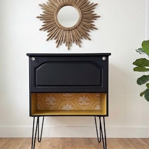 May include: A black cabinet with a drop-down door, two clear knobs, and four black metal hairpin legs. The open lower shelf features a mustard yellow interior with a white floral pattern. Above the cabinet hangs a gold sunburst wall mirror.