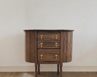May include: A dark brown wooden cabinet featuring three stacked drawers with rounded sides and metallic copper-toned hardware. The piece stands on four slender, turned legs and has a smooth, polished finish that highlights the natural wood grain.