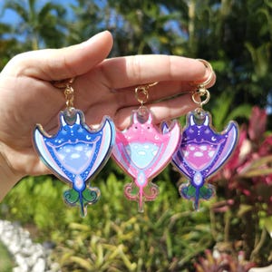 May include: Three iridescent keychains featuring a stylised manta ray design. Each keychain is a different colour: blue, pink and purple. The manta rays have a white and blue cloud pattern on their bodies.