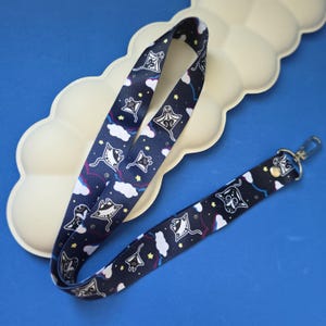May include: A navy blue lanyard with a repeating pattern of white manta rays, clouds, and stars. It has a metal clasp and swivel hook. The background is a light blue surface.