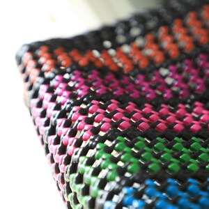 May include: Close-up of a colorful woven mat with black, pink, green, blue, and orange stripes. The mat is made of a thick, durable material and has a textured surface.