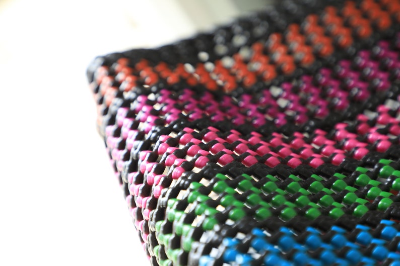 May include: Close-up of a colorful woven mat with black, pink, green, blue, and orange stripes. The mat is made of a thick, durable material and has a textured surface.