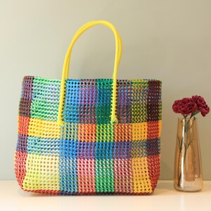 May include: A colorful checkered tote bag with yellow handles. The bag is made of woven plastic and has a bright, cheerful design.