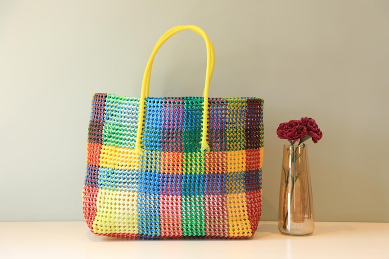 May include: A colorful checkered tote bag with yellow handles. The bag is made of woven plastic and has a bright, cheerful design.
