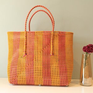 May include: A yellow and pink striped woven tote bag with thick handles. The bag is made of a plastic material and has a woven pattern.