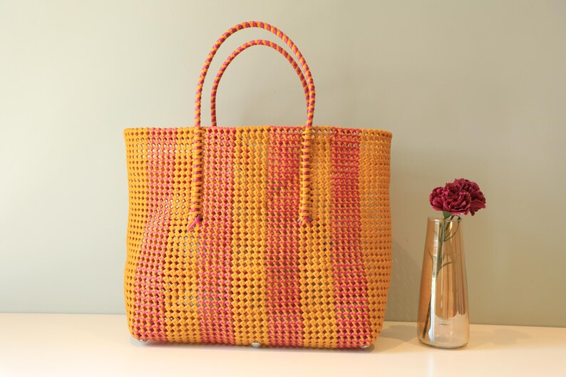 May include: A yellow and pink striped woven tote bag with thick handles. The bag is made of a plastic material and has a woven pattern.