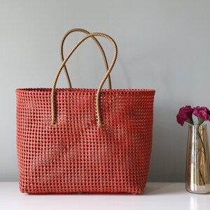 May include: A red woven tote bag with brown leather handles. The bag is made of a woven plastic material and has a rectangular shape.
