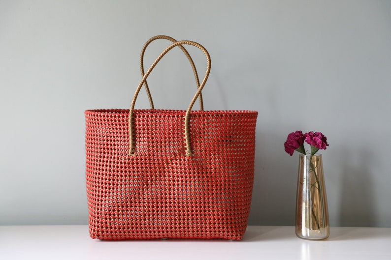 May include: A red woven tote bag with brown leather handles. The bag is made of a woven plastic material and has a rectangular shape.