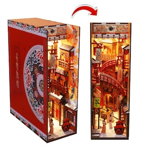 Puede incluir: Un rincón de libro rojo e iluminado que representa una detallada escena callejera asiática en miniatura. El rincón del libro tiene una cubierta roja decorativa con un diseño floral. El interior muestra una calle de varios niveles con puentes y figuras.