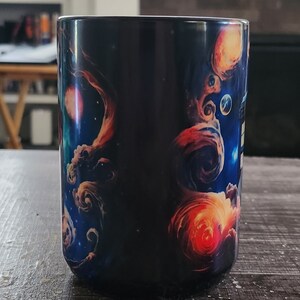 Op de afbeelding: Een zwarte keramische mok met een kleurrijk galaxy-ontwerp. Het ontwerp bevat draaiende nevels, sterren en planeten in tinten blauw, oranje en rood.
