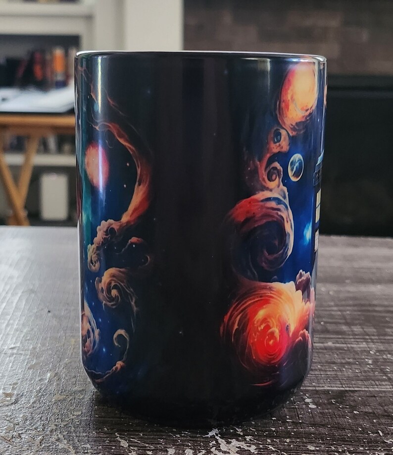 Op de afbeelding: Een zwarte keramische mok met een kleurrijk galaxy-ontwerp. Het ontwerp bevat draaiende nevels, sterren en planeten in tinten blauw, oranje en rood.