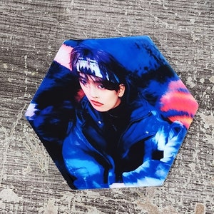 May include: A close-up of a person wearing a blue jacket with a black bandana. The person has dark hair and is looking at the camera. The background is a blurry blue and pink.