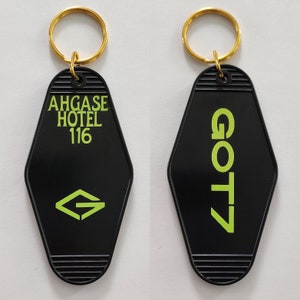 Puede incluir: Dos llaveros negros con forma de llaves de habitación de motel. Un llavero tiene el texto "AHGASE HOTEL 116" y una letra estilizada "G" en verde. El otro llavero tiene el texto "GOT7" en verde.