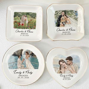 May include: Four white ceramic trinket dishes with gold-trimmed edges. Two are square, one is circular, and one is heart-shaped. Each features a custom illustration of a couple with names and dates: Charlie & Anna Married 27.08.2026 and Emily & Andy Engaged 26.05.2026.