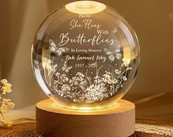 May include: A clear glass sphere with the text "Now She Flies With Butterflies" and "In Loving Memory Bob Samuel Irby 1937-2026". The sphere sits on a round wooden base, illuminated from within. Butterflies and floral designs are etched inside.
