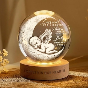 May include: A glass sphere featuring an etched angel on a crescent moon, set on a wooden base. The sphere displays the text "Carried for a moment, Loved for a lifetime, Baby Taneira - 2026." The base reads "Forever in our hearts."