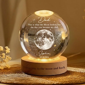 May include: A clear glass sphere with a detailed moon image inside, resting on a wooden base. The sphere has the text "Dad" and "Sophia 05.20.2026" engraved on it. The base reads "I love you to the moon and back."