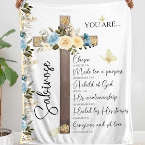 May include: A white fleece blanket featuring a cross design with floral embellishments. The cross is crafted from wood and has a gold trim. The blanket displays the text "You Are..." and a list of biblical verses about being chosen, made for a purpose, a child of God, His workmanship, healed by His stripes, forgiven and set free. The name "Sabirose" is written vertically on the left side of the blanket.