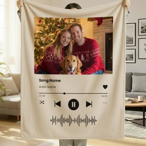 May include: A cream-coloured blanket with a framed photograph of a couple and a dog, overlaid with a music player interface. The photo shows a couple in red sweaters in front of a Christmas tree. The blanket has the text "Song Name" and "Artist Name".