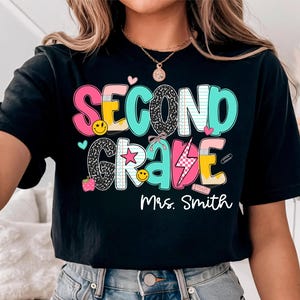 May include: Black t-shirt with the words "SECOND GRADE" and "Mrs. Smith" in colorful, playful lettering. The design includes hearts, stars, smiley faces, and school-themed graphics. The shirt is a comfortable, casual style.