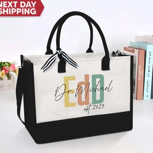 May include: A black and white tote bag with a personalized design. The bag has a black canvas bottom and a white canvas top with the text "Dr. Michael EDD est. 2025" in colorful letters.
