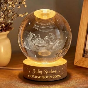 May include: A glowing crystal ball lamp with an ultrasound image of a baby inside. The lamp sits on a wooden base engraved with "Baby Sophia COMING SOON 2026". The lamp is lit from within, creating a warm, inviting glow.