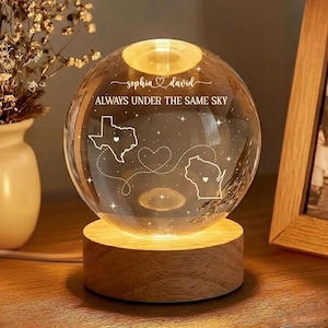 May include: A glowing crystal ball night light with a wooden base. The sphere is engraved with the names "Sophia & David", the phrase "Always Under The Same Sky", and outlines of two states connected by a dotted line. The light is warm and inviting.