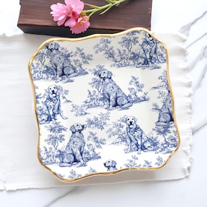 Può includere: Un piatto da portata quadrato in ceramica bianca con un motivo toile de Jouy blu e bianco a tema cani. Il piatto ha un bordo smerlato con finiture dorate. Il motivo presenta cani in un ambiente paesaggistico.